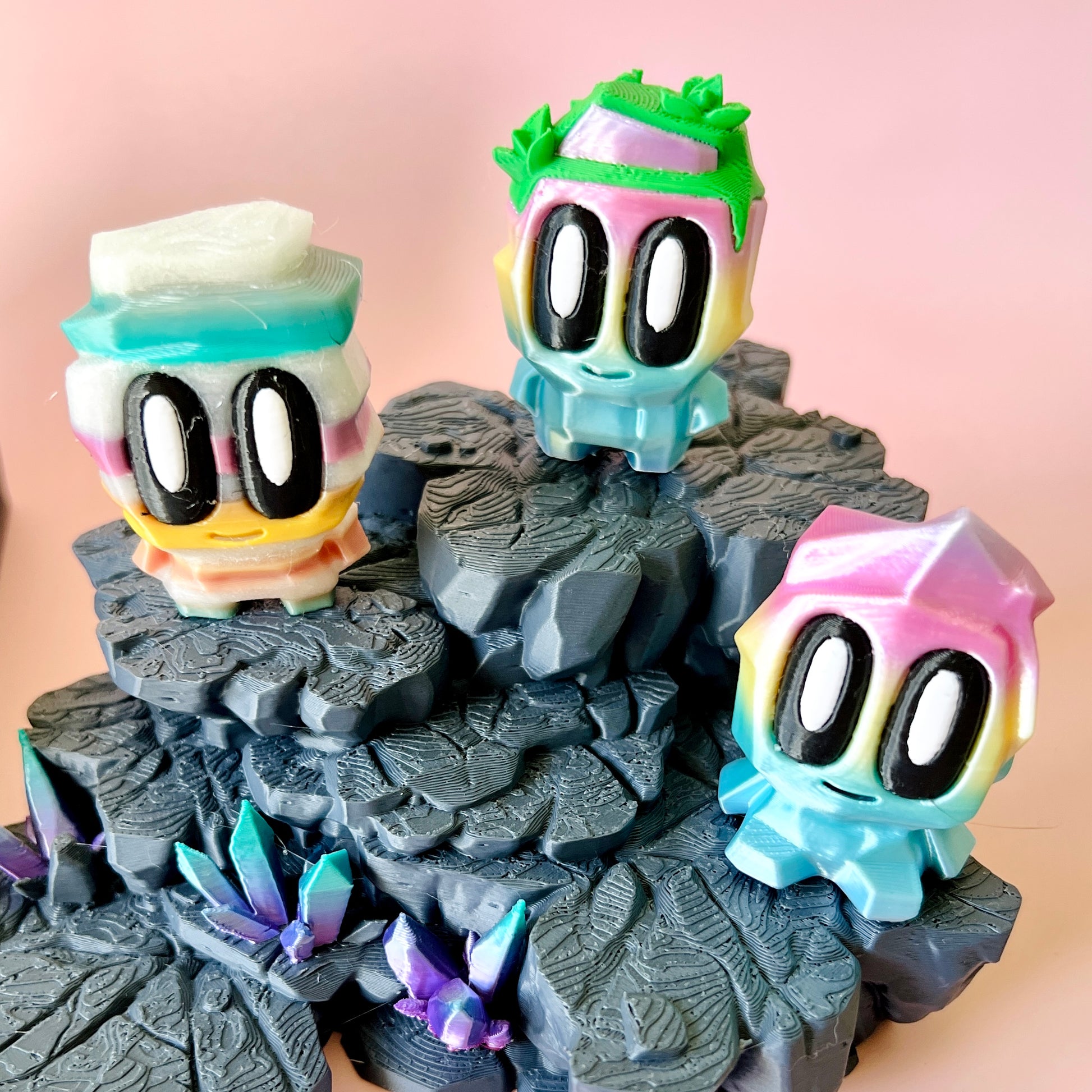 Three colorful, cartoonish figures with large eyes stand on textured gray rocks, surrounded by shimmering, crystal-like forma
