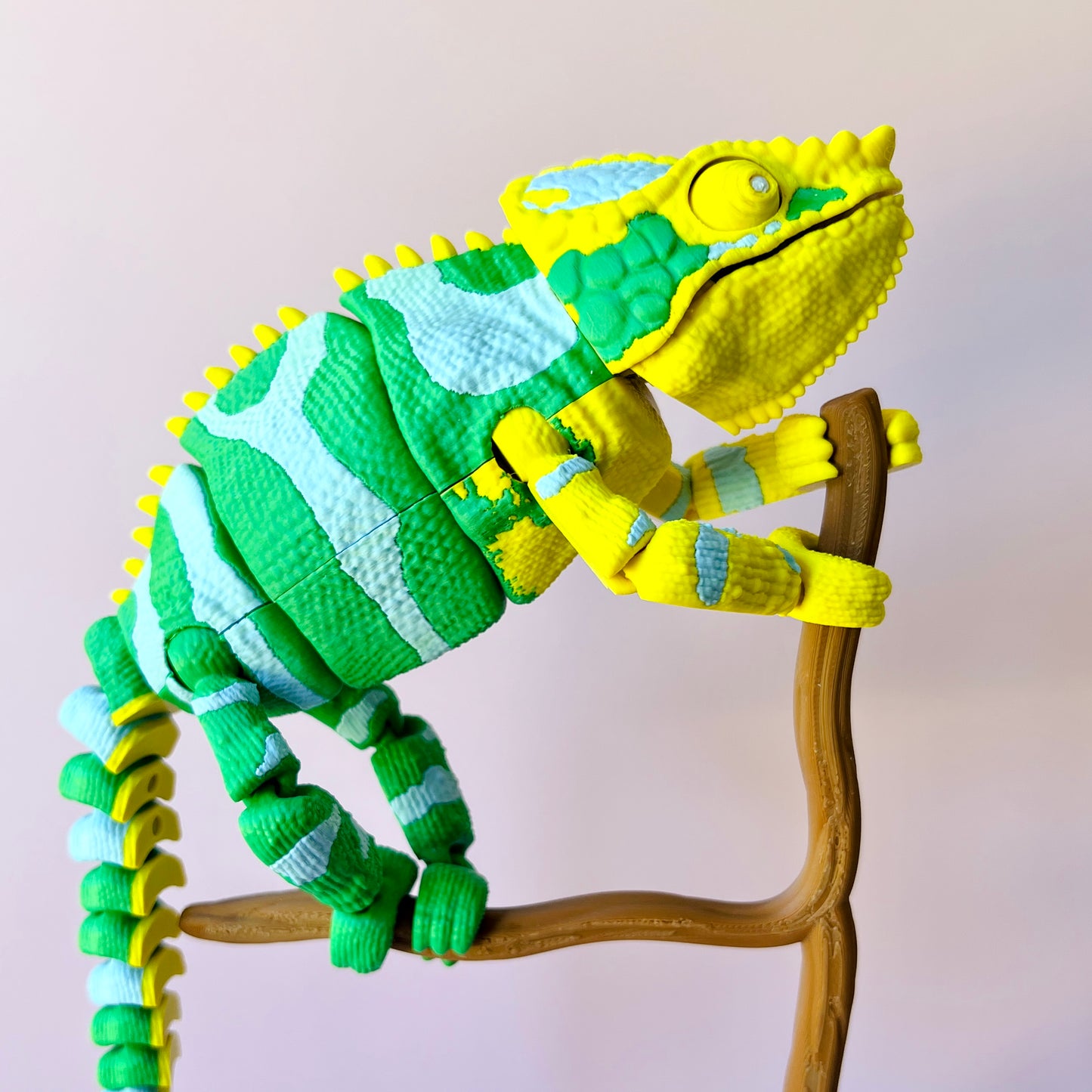 Colorful chameleon figure with green and yellow patterns climbing a brown branch against a soft pink background.