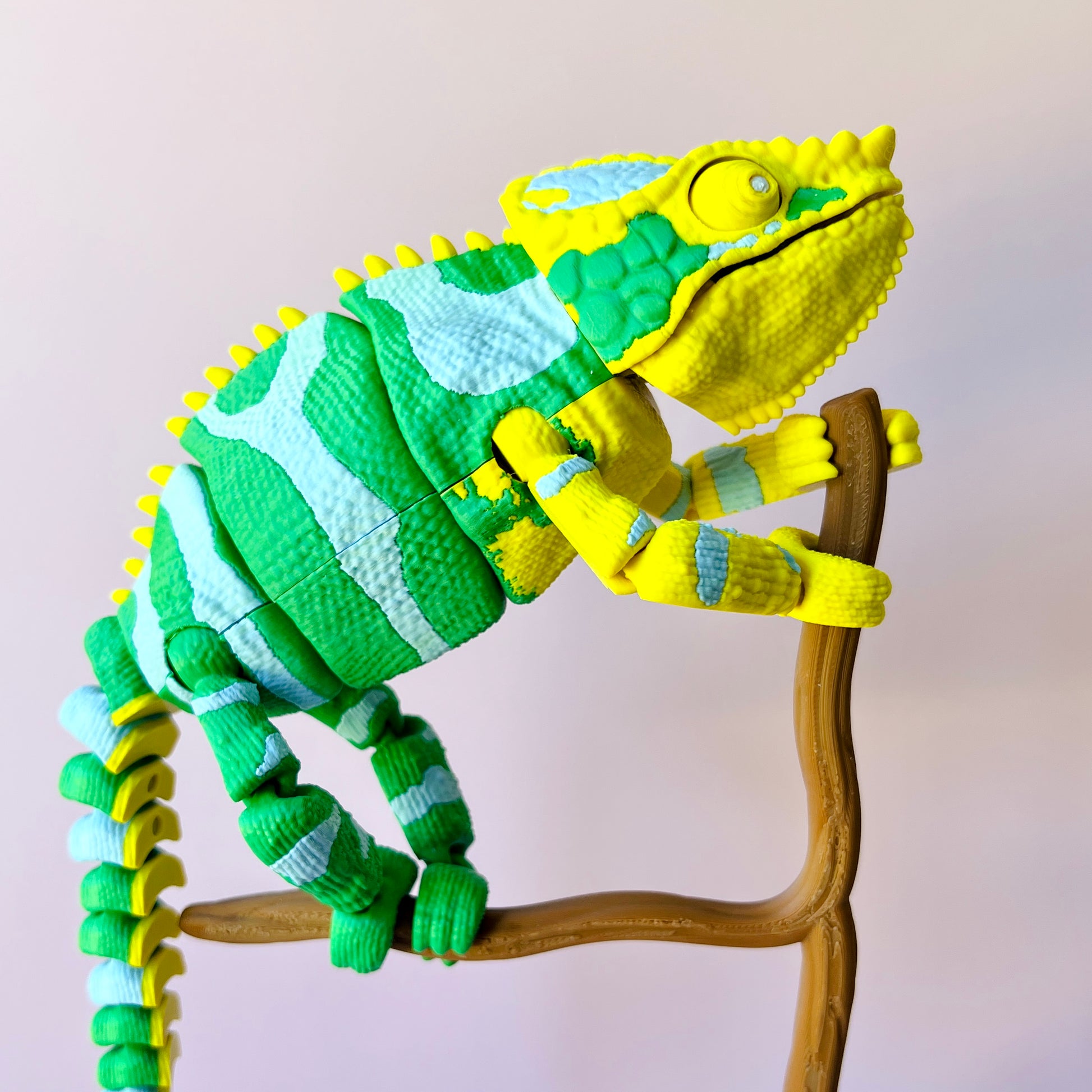 Colorful chameleon figure with green and yellow patterns climbing a brown branch against a soft pink background.