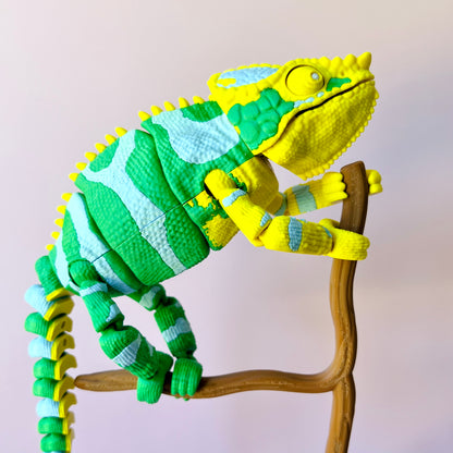 Colorful chameleon figure with green and yellow patterns climbing a brown branch against a soft pink background.