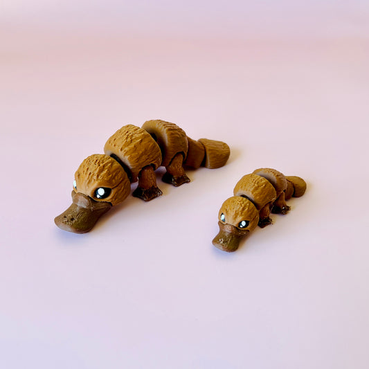 Two stylized platypus figures, one larger and one smaller, with detailed textures and bright eyes against a soft pink backgro