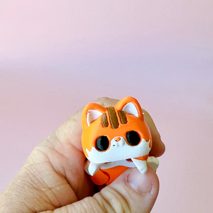 A bright orange cat figure with large eyes and a cute expression, held in a hand against a soft pink background.