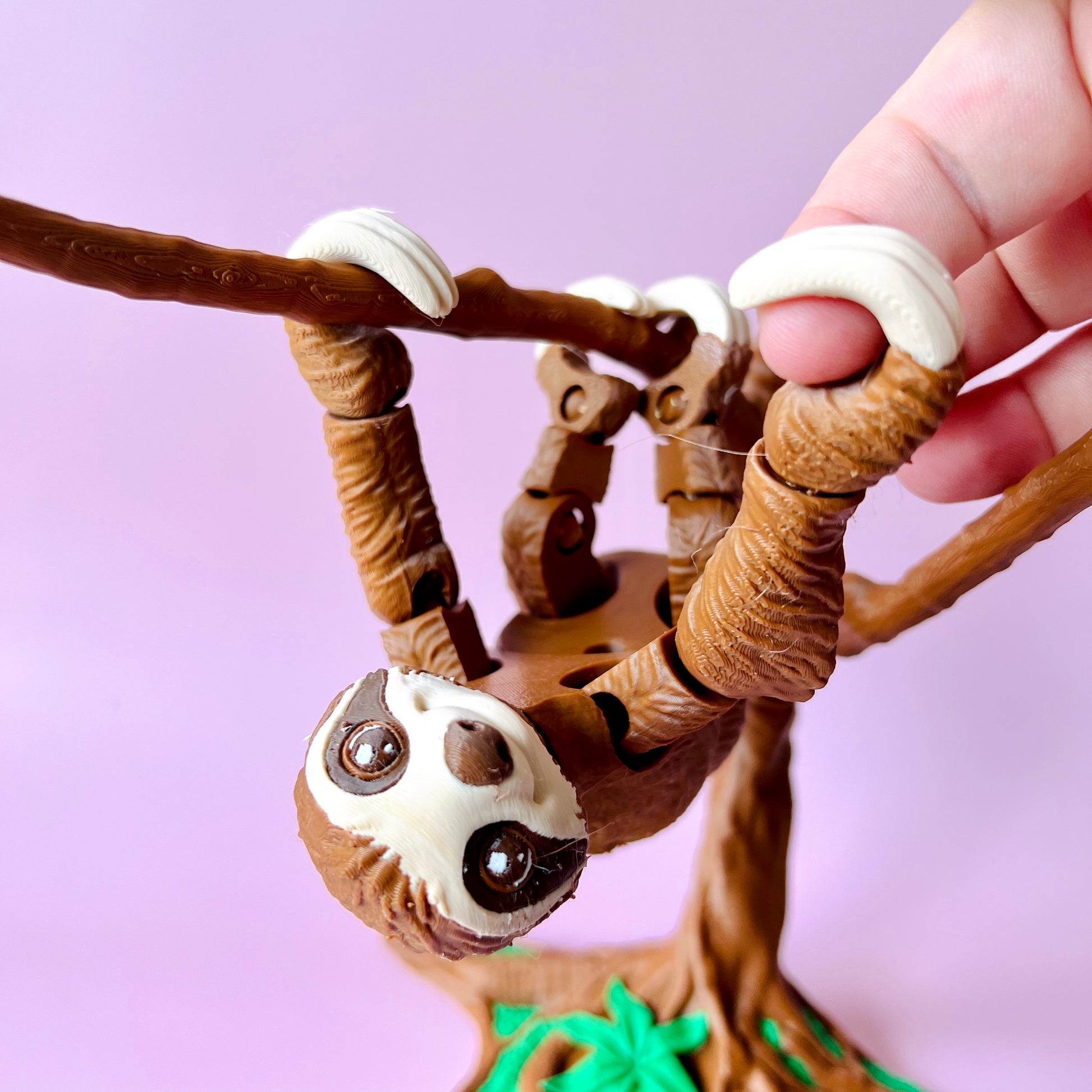 A sloth hangs upside down from a branch, with large eyes and detailed fur, against a soft pink background.