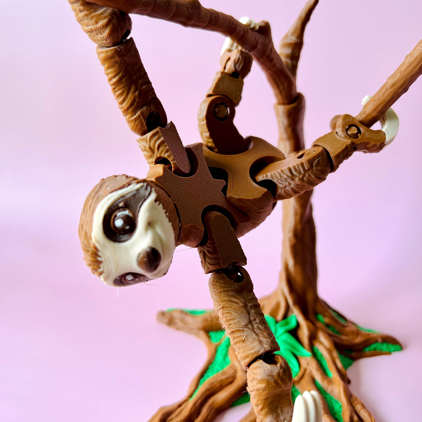 A sloth hangs from a branch, featuring movable limbs and a playful expression against a soft pink background.