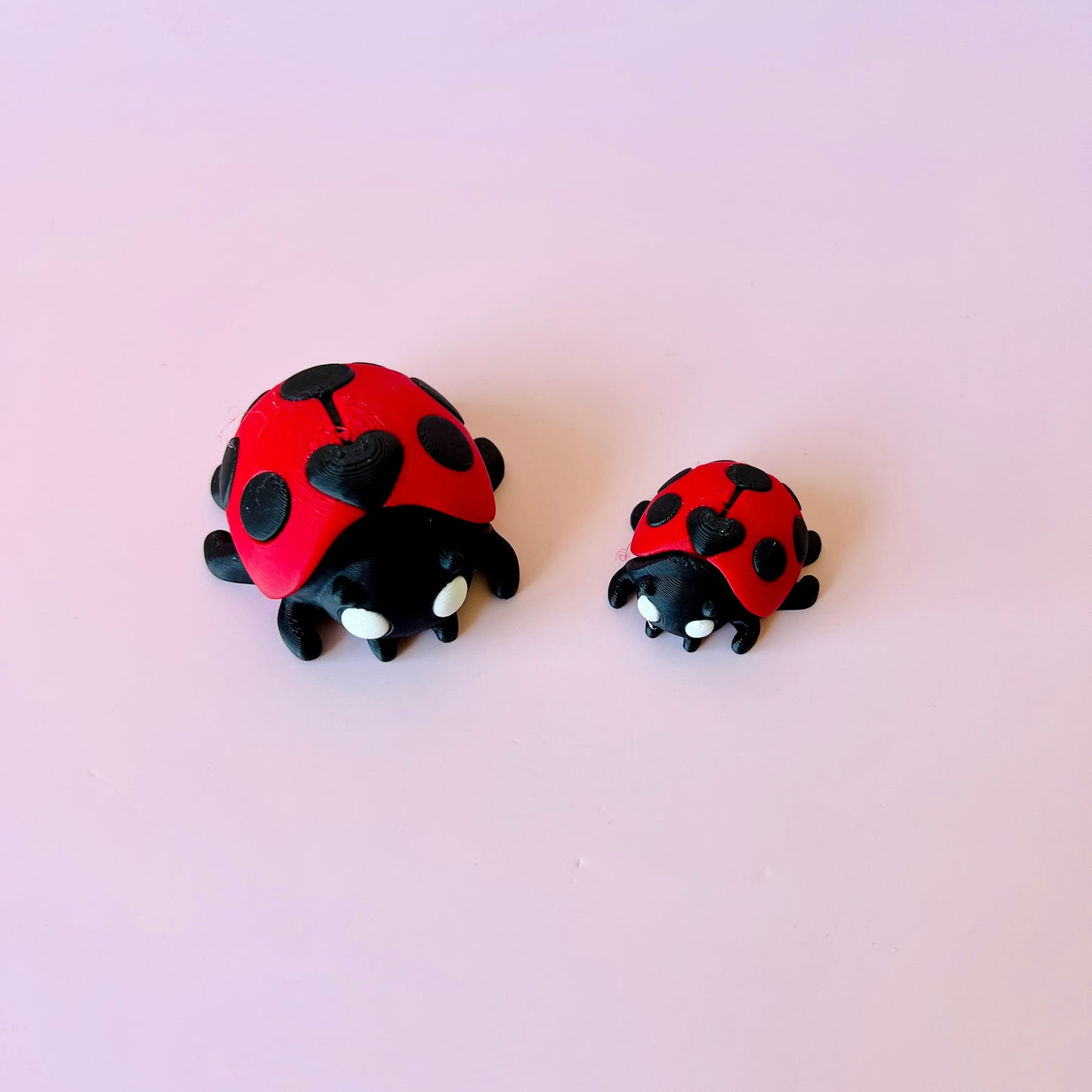 Two stylized ladybugs, one larger and one smaller, with red shells and black spots, against a soft pink background.