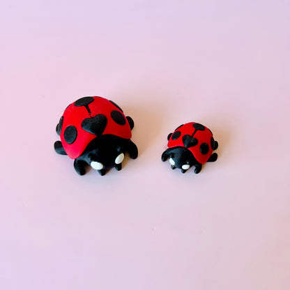 Two stylized ladybugs, one larger and one smaller, with red shells and black spots, against a soft pink background.