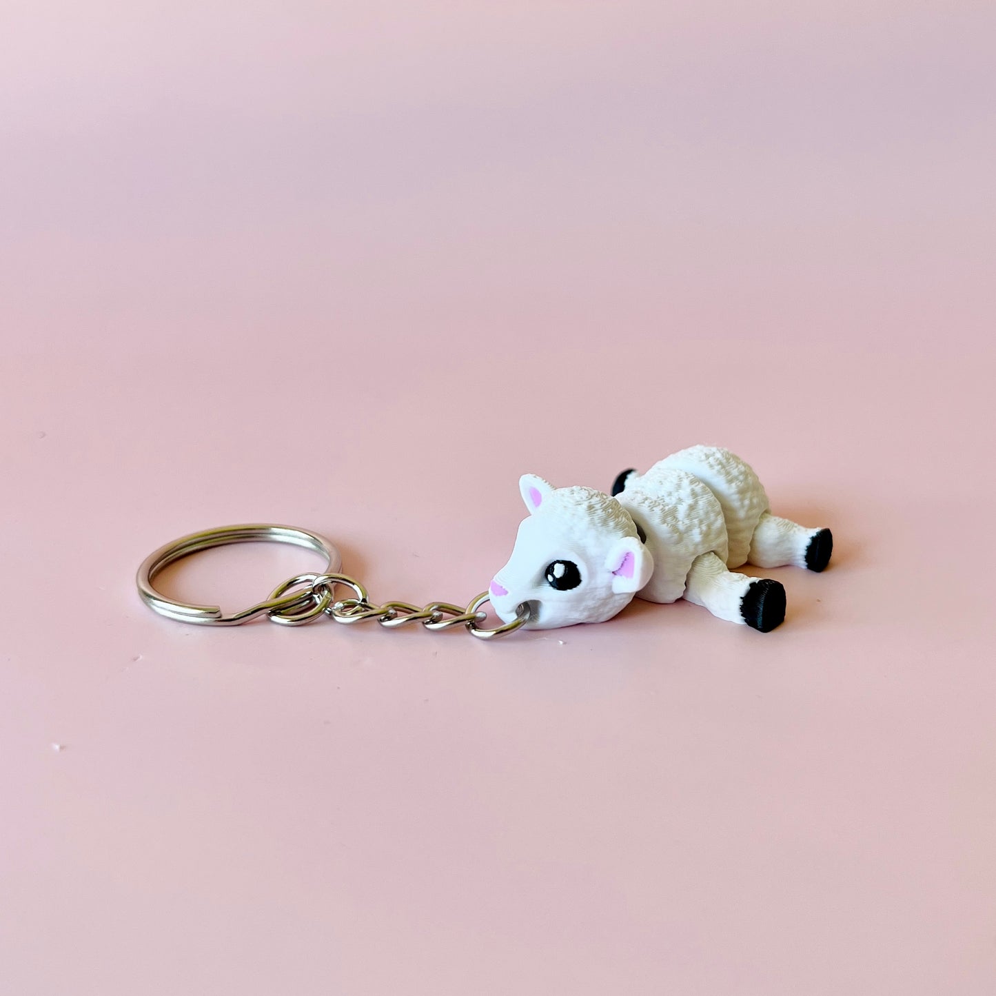 A cute white sheep charm with big eyes, attached to a silver keychain, set against a soft pink background.