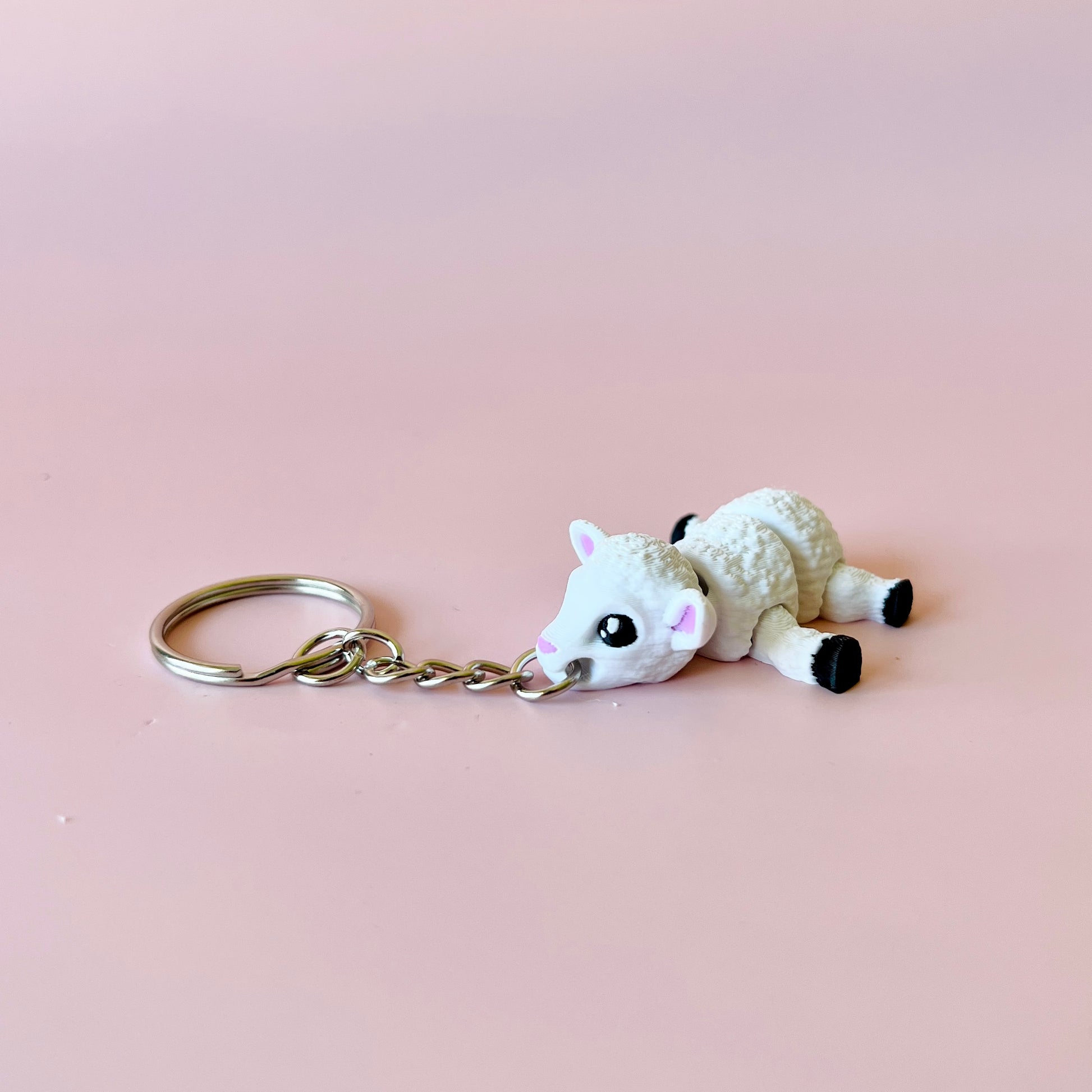 A cute white sheep charm with big eyes, attached to a silver keychain, set against a soft pink background.