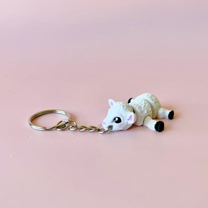A cute white sheep charm with big eyes, attached to a silver keychain, set against a soft pink background.
