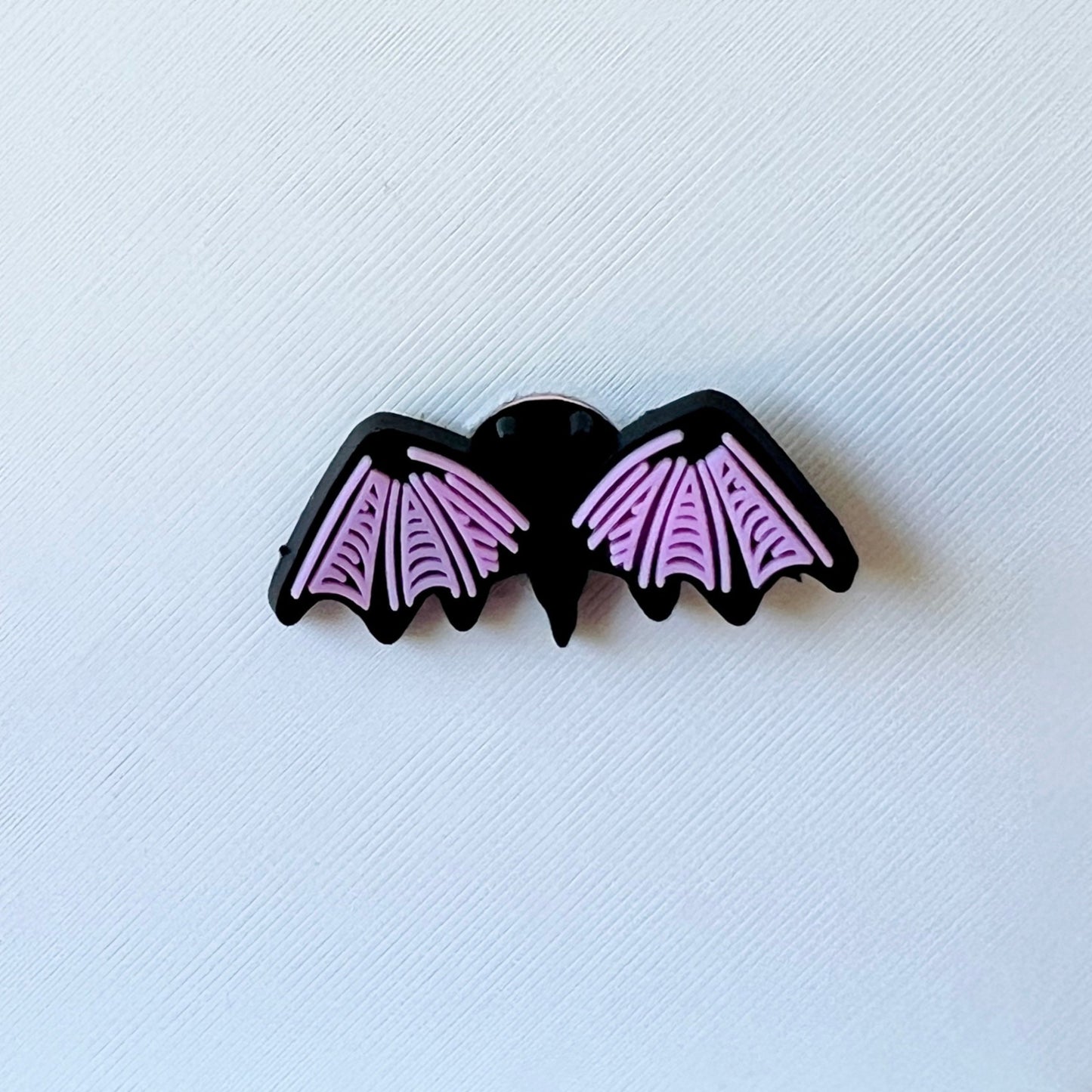 A black bat with pink wings, featuring detailed patterns, rests on a textured white background.