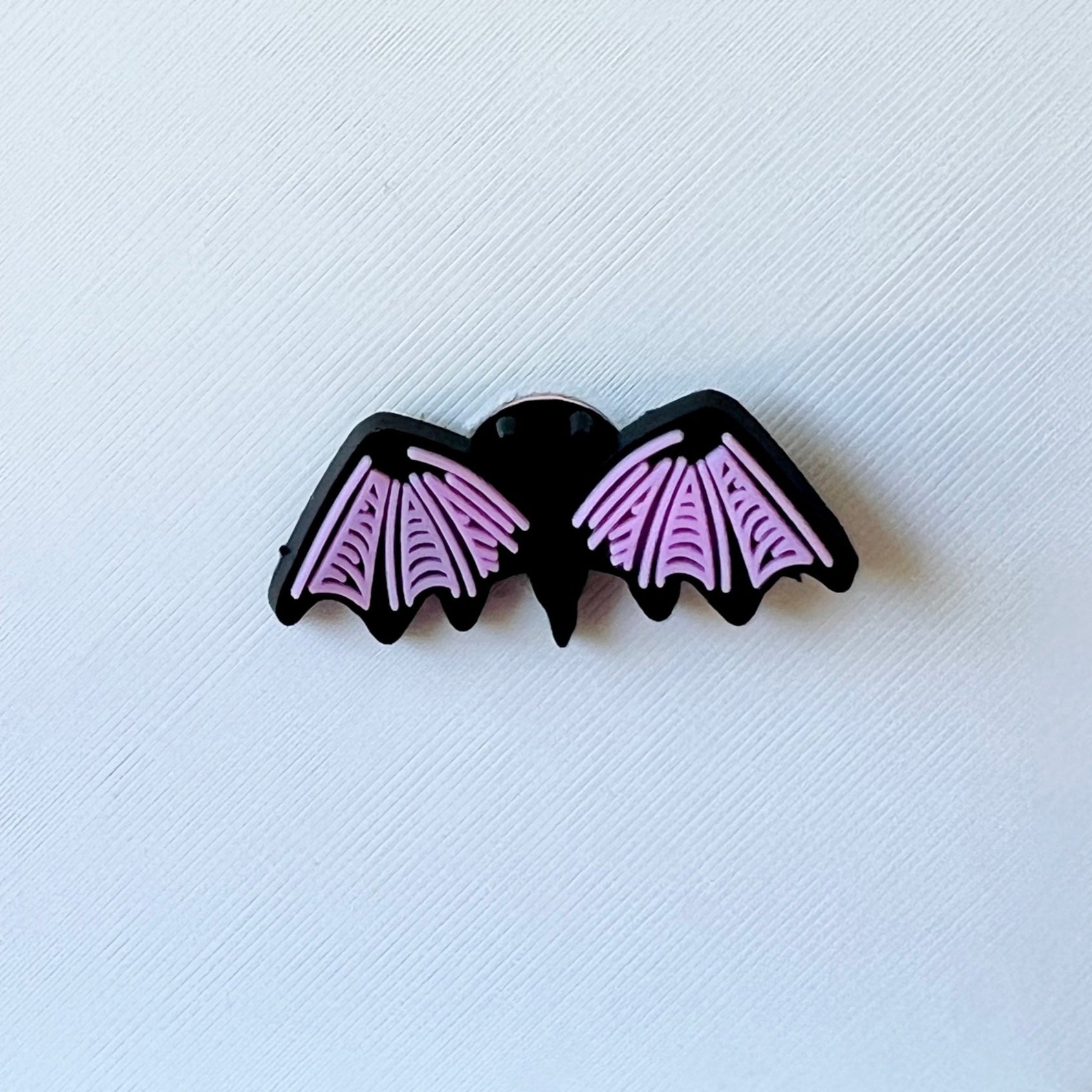 A black bat with pink wings, featuring detailed patterns, rests on a textured white background.