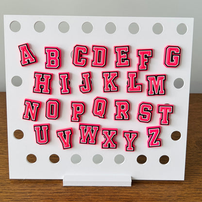 Colorful pink alphabet charms with bold white and black outlines displayed on a white board with circular holes.