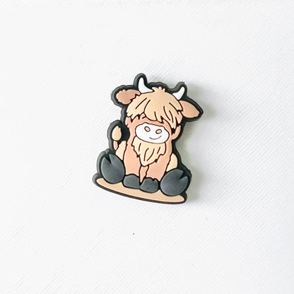 Adorable sitting Highland cow charm with a fluffy coat and smiling face, perfect for adding charm to your footwear.