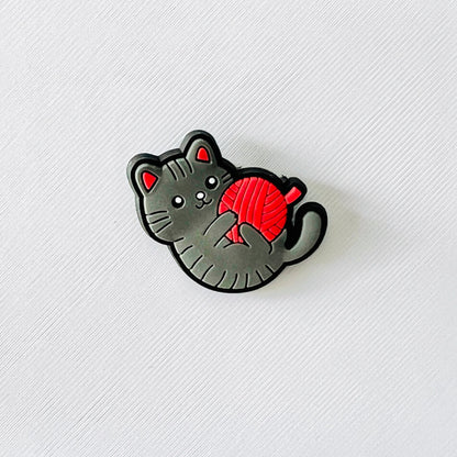 Playful grey cat charm tangled in a bright red ball, perfect for adding a whimsical touch to your favorite footwear.