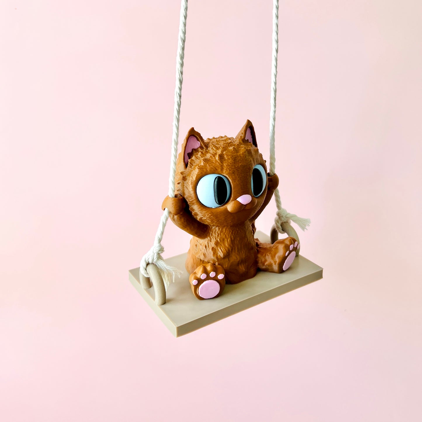 Adorable 3D-printed cat figurine on a swing, featuring large expressive eyes and tiny paws, against a soft pink background.