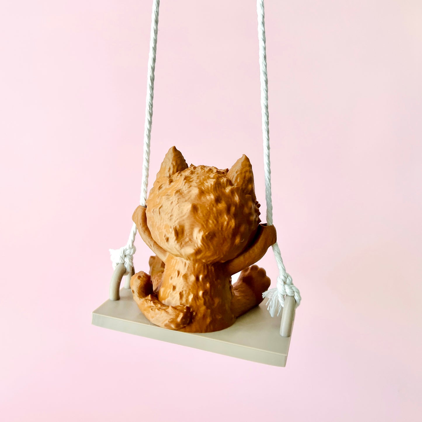 Adorable 3D-printed cat figurine on a swing, showcasing playful details and expressive features against a soft pink backgroun