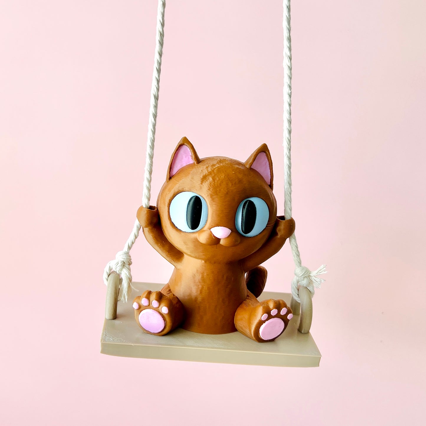 Adorable 3D-printed cat figurine on a swing, featuring big expressive eyes and tiny paws, set against a soft pink background.