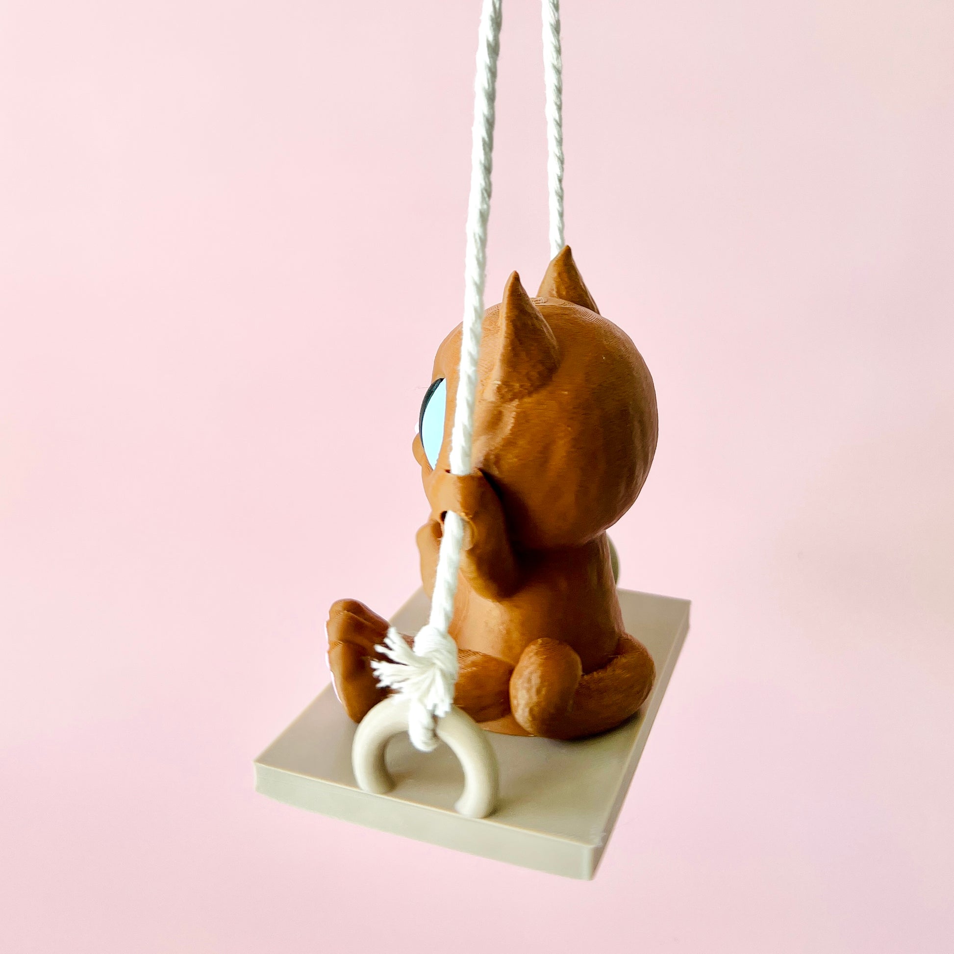 Adorable 3D-printed cat figurine swinging joyfully, showcasing charming details and expressive eyes against a soft pink backg