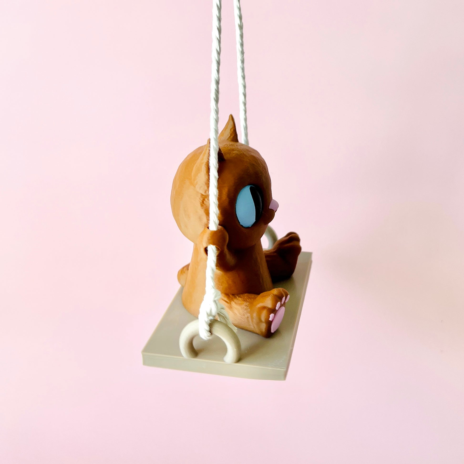 Adorable 3D-printed cat figurine sitting on a swing, with big expressive eyes and tiny paws gripping the ropes.
