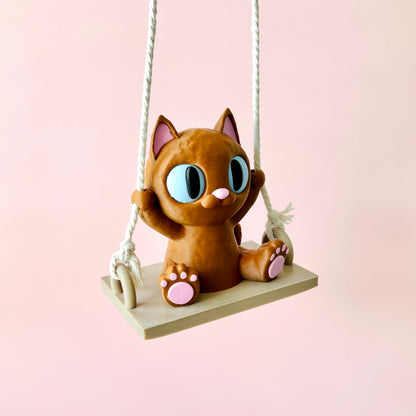 Adorable 3D-printed cat figurine on a swing, featuring expressive eyes and tiny paws, against a soft pink background.