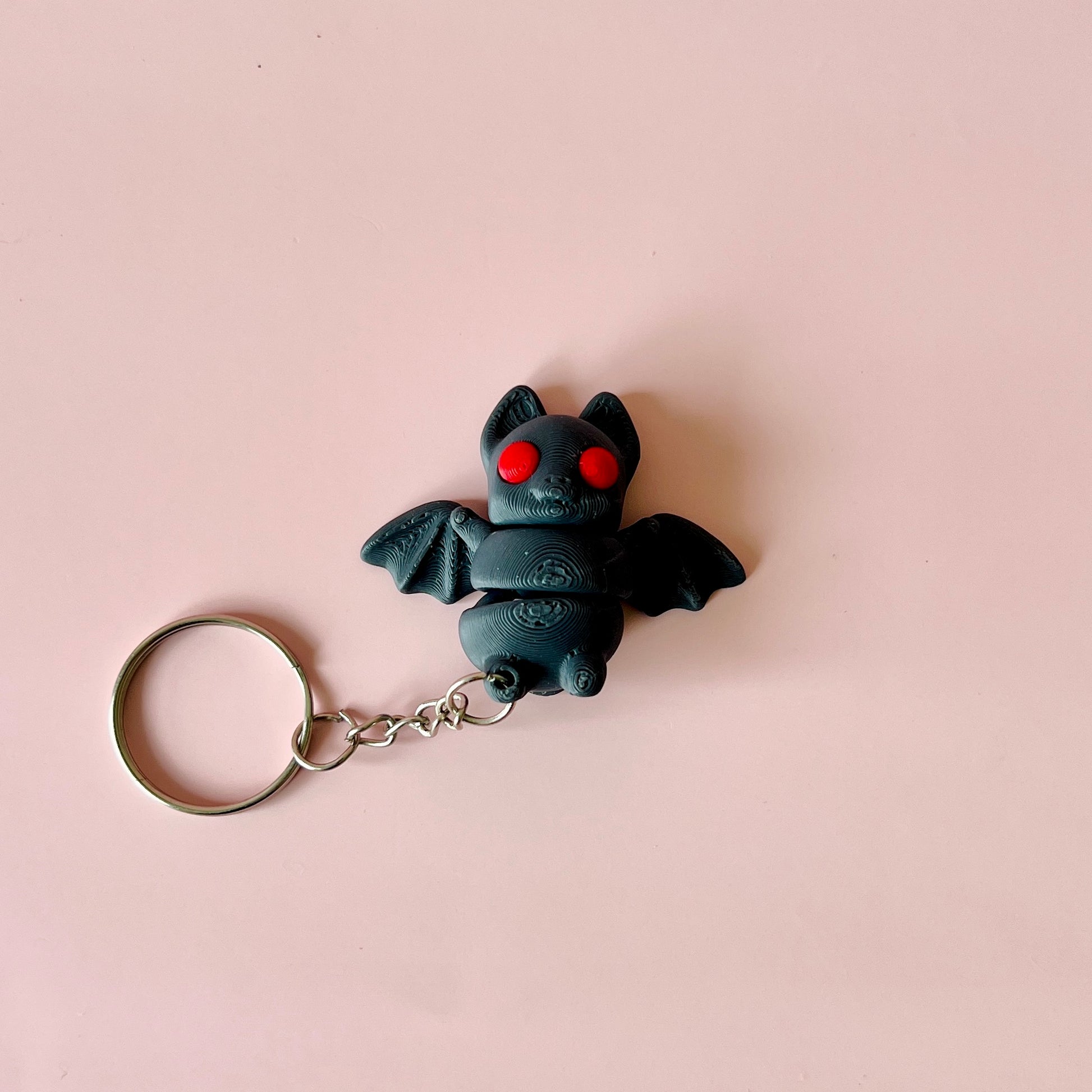 A small black bat figure with red eyes, attached to a silver keychain, against a soft pink background.