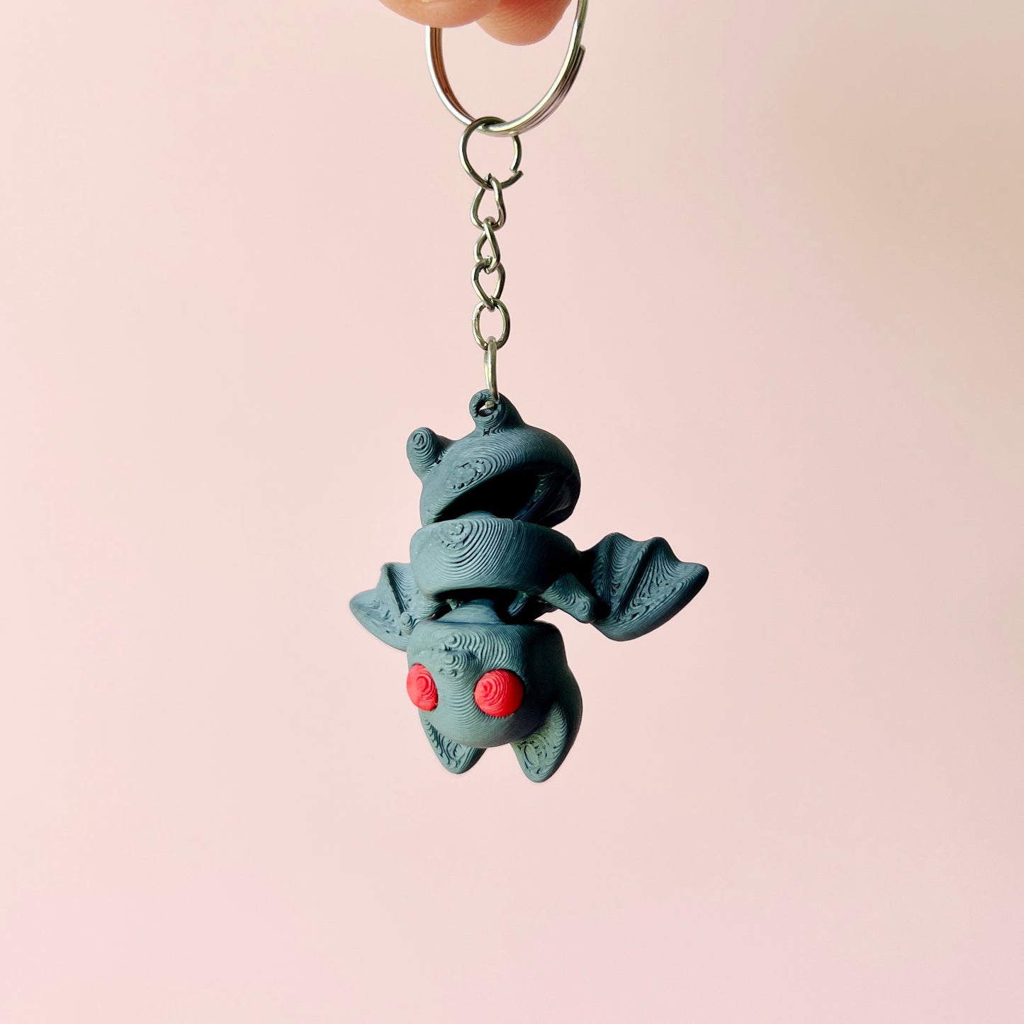 A whimsical, gray creature with red eyes hangs from a keychain against a soft pink background.
