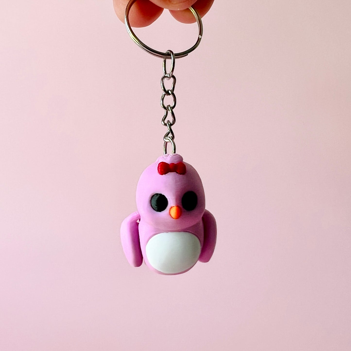 A cute pink bird keychain with black eyes and a red bow, suspended against a soft pink background.