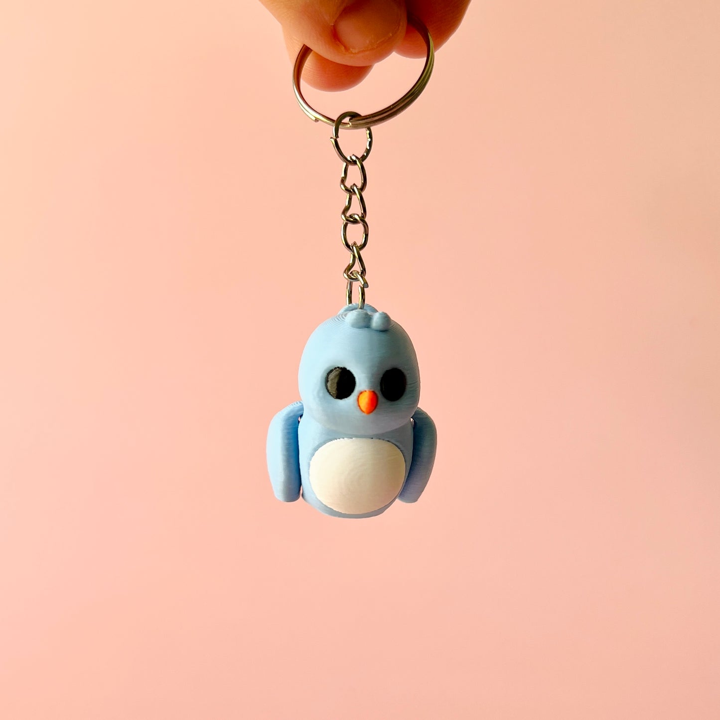 A cute blue bird figure with a round white belly, hanging from a keychain against a soft pink background.