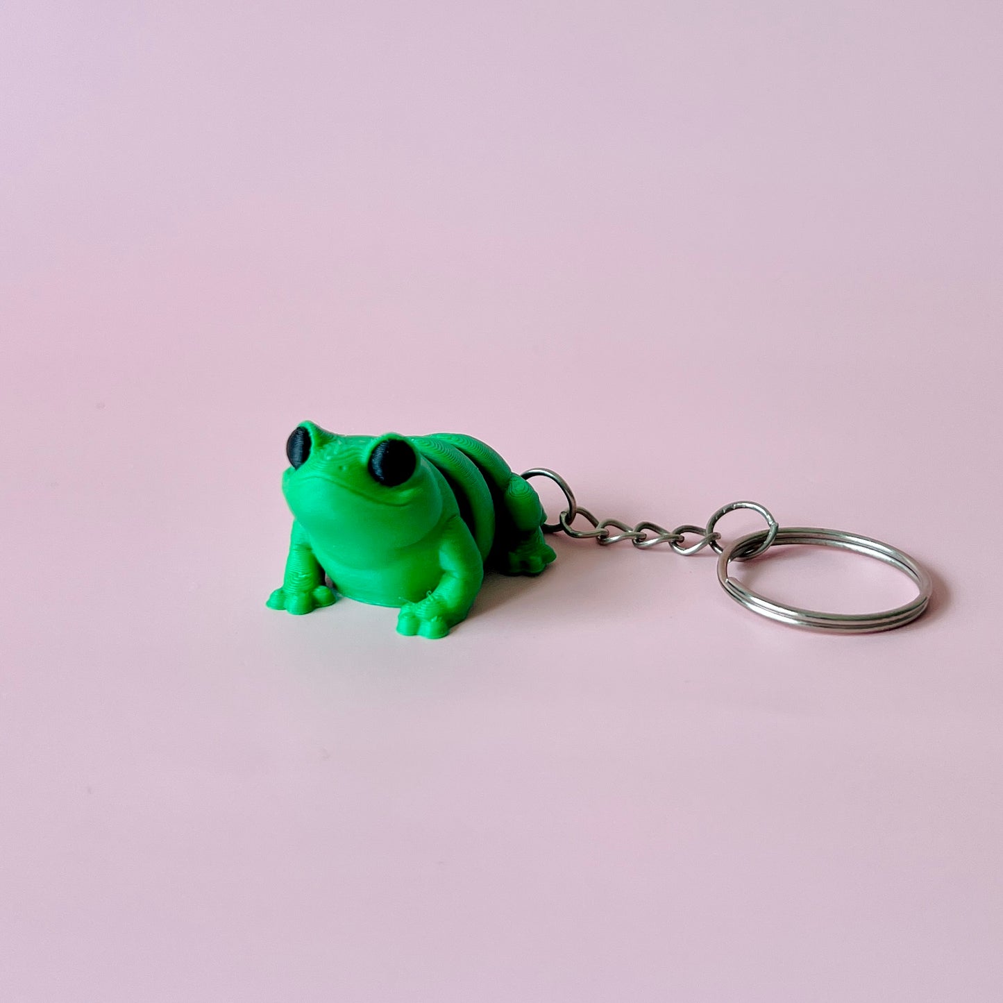 Bright green frog figure with large black eyes, attached to a metal keyring, set against a soft pink background.
