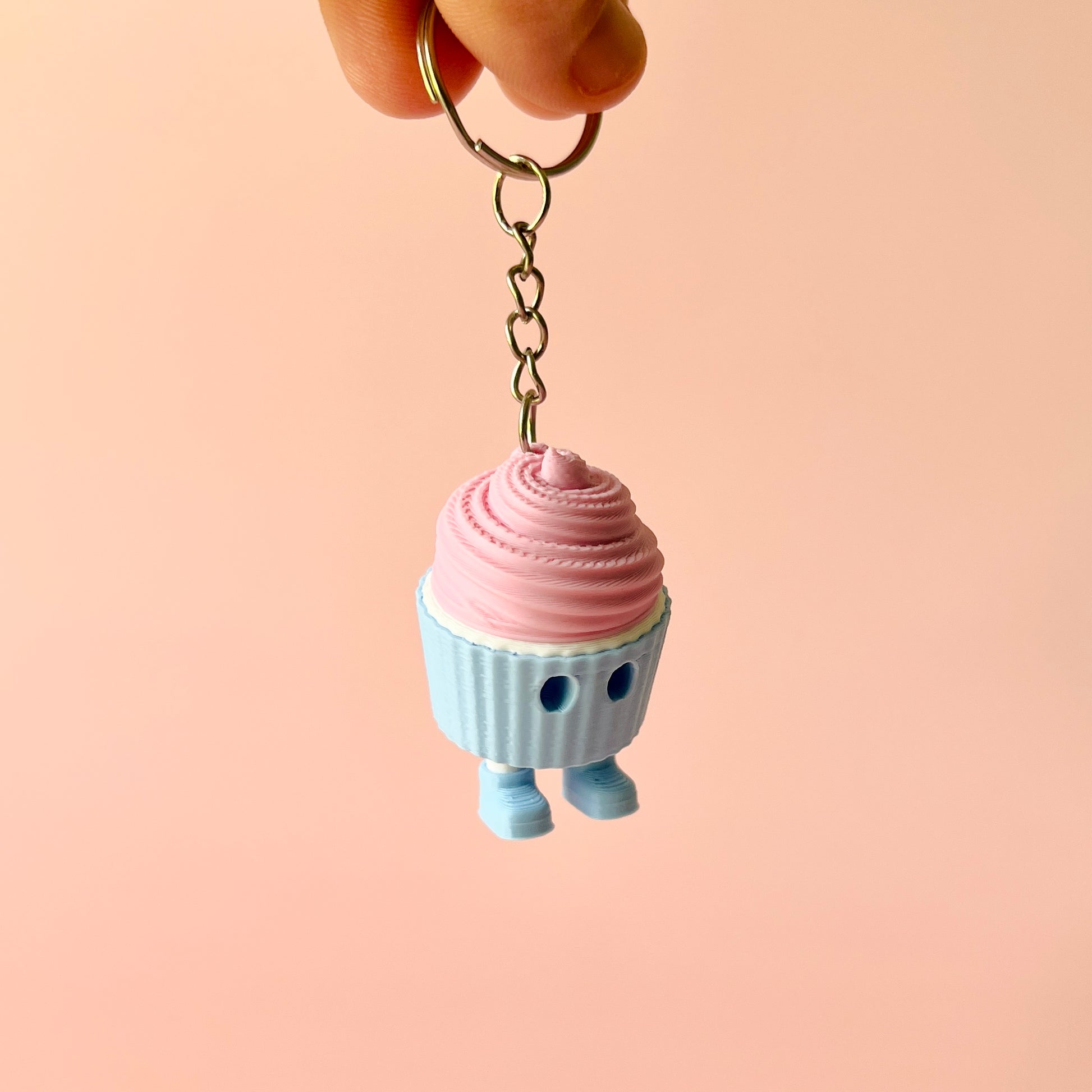 A hand holds a colorful cupcake keychain against a soft pink background, showcasing whimsical details and charm.