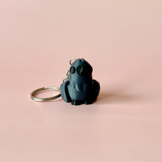 A small dark bird figurine with a keyring, set against a soft pink background.