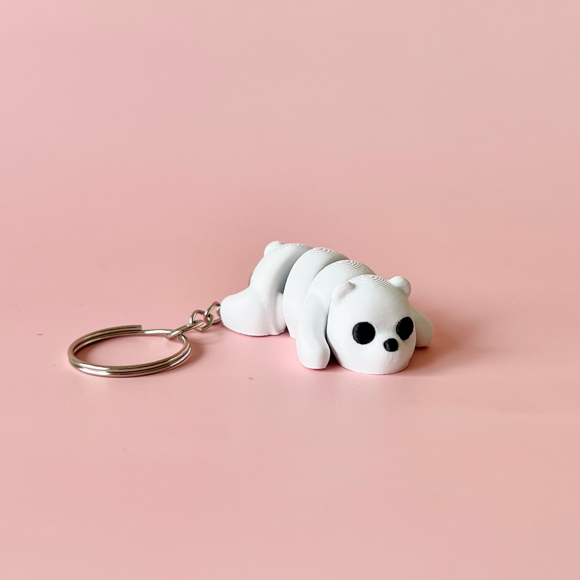 A small white figure of a bear lying down, with black eyes and a silver keyring, set against a soft pink background.