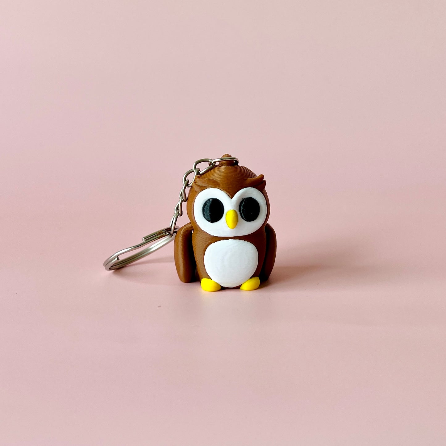 A cute brown owl figure with big eyes, a yellow beak, and tiny feet, attached to a metal keychain on a pink background.