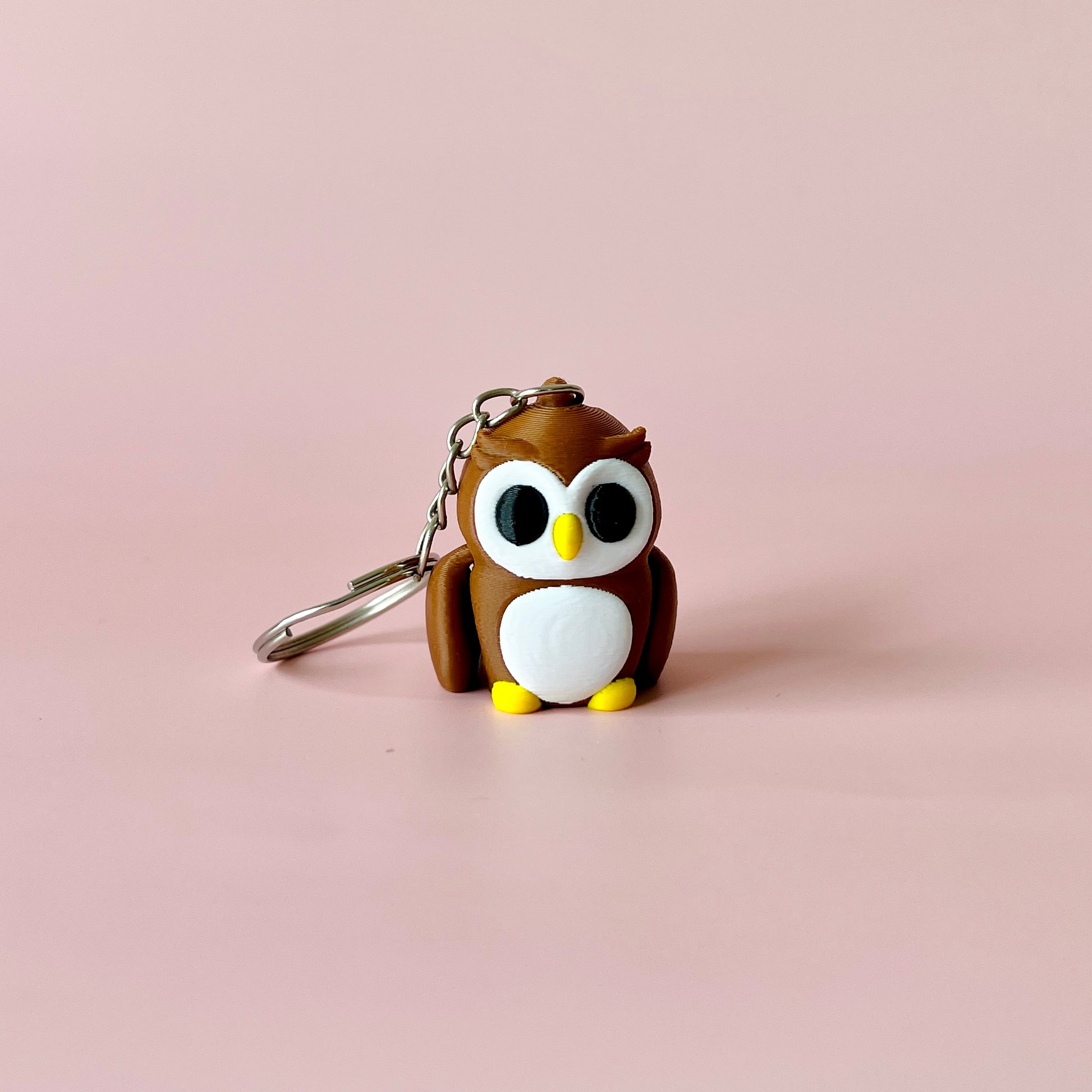 A cute brown owl figure with big eyes, a yellow beak, and tiny feet, attached to a metal keychain on a pink background.