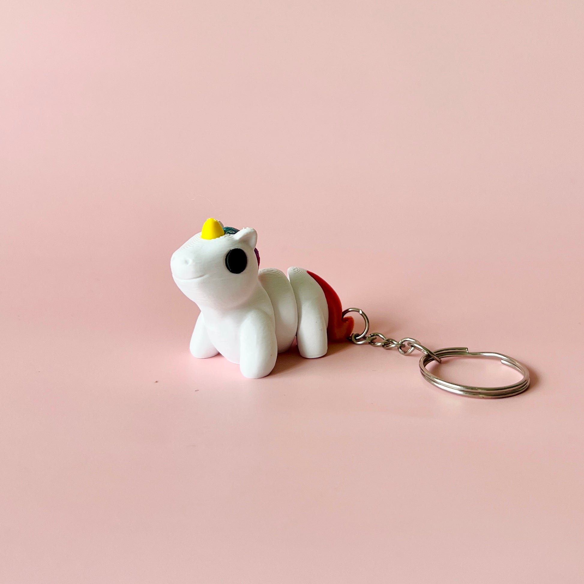 A small, cheerful unicorn figurine with a yellow horn and rainbow tail on a soft pink background.