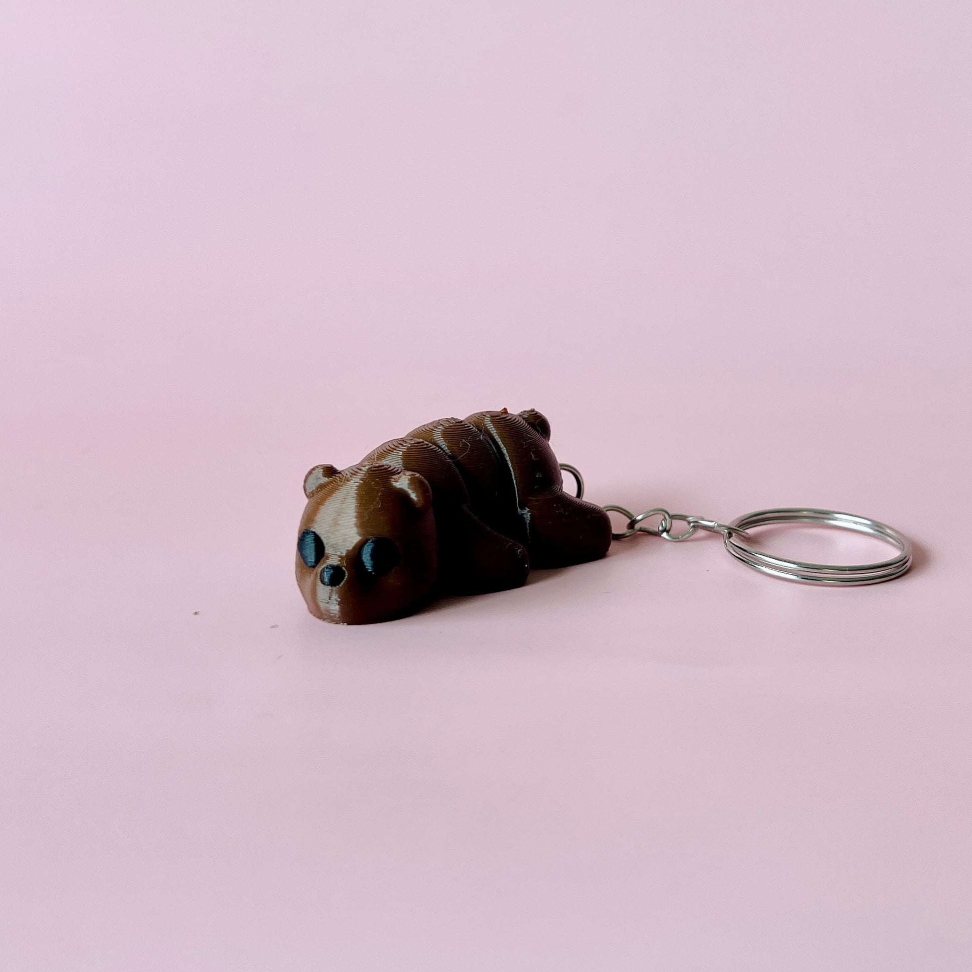A cute brown bear figure with black eyes rests on a soft pink background, attached to a silver keychain.