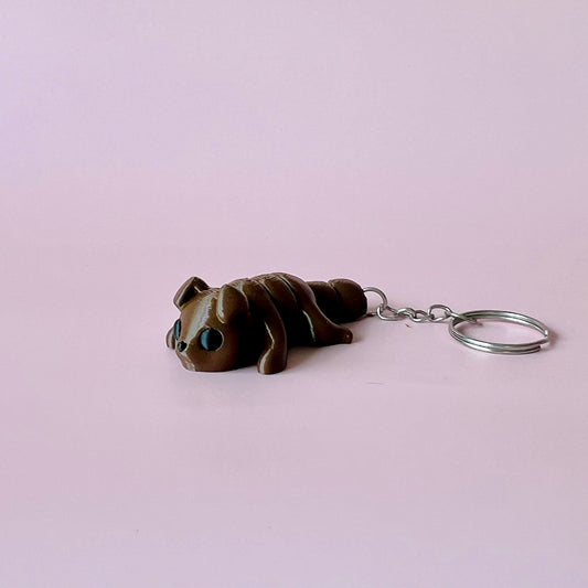 A cute brown creature with black eyes, resting on a soft pink background, attached to a metal keyring.