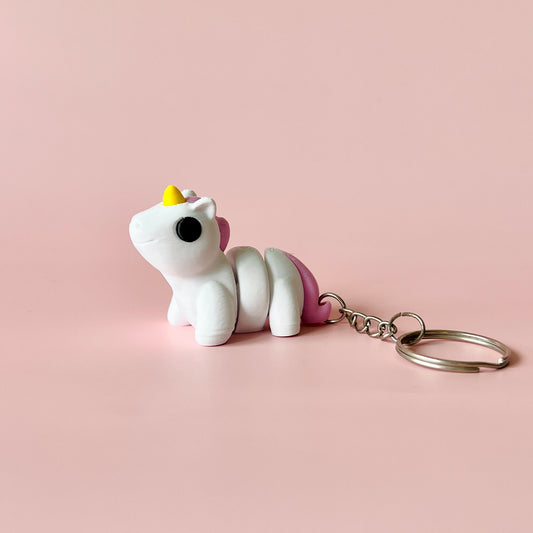 A whimsical white unicorn keychain with a pink mane and yellow horn, set against a soft pink background.