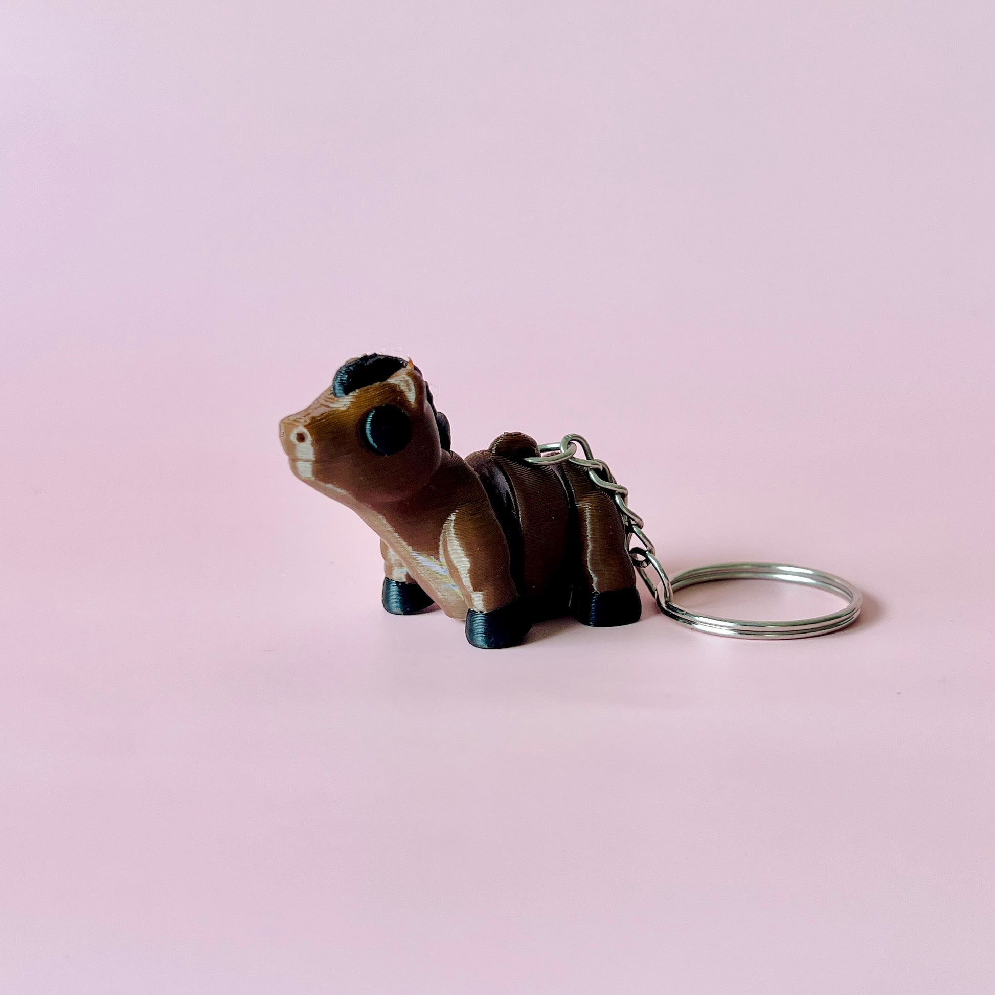 A small brown figurine of a horse with black accents, attached to a metal keyring against a soft pink background.