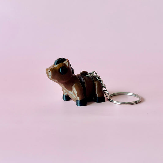 A small brown figurine of a horse with black accents, attached to a metal keyring against a soft pink background.