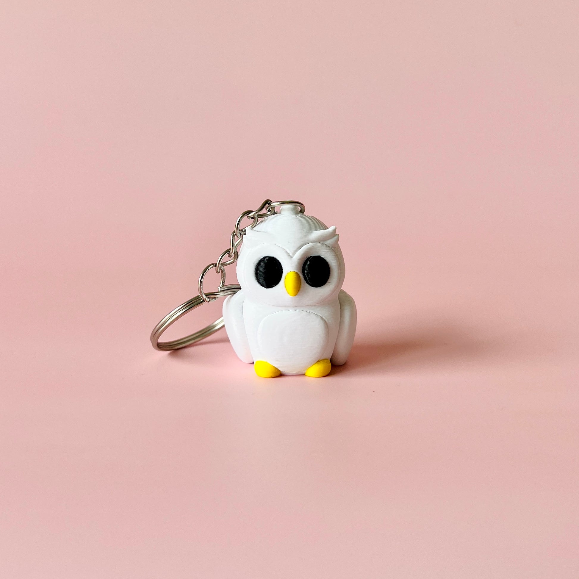 A cute white owl figure with large black eyes and yellow feet, attached to a silver keychain against a pink background.