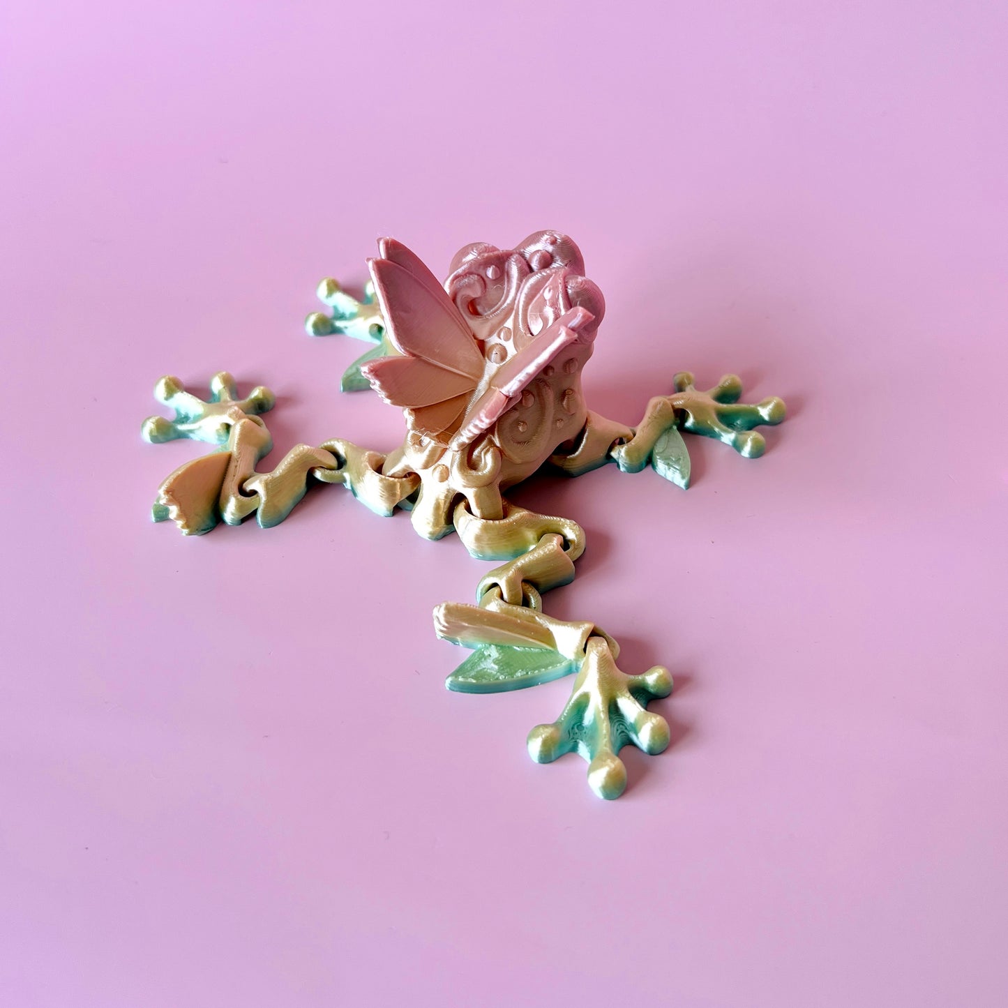 Whimsical butterfly frog figurine with delicate details, showcasing a blend of vibrant colors and charm on a soft pink backgr