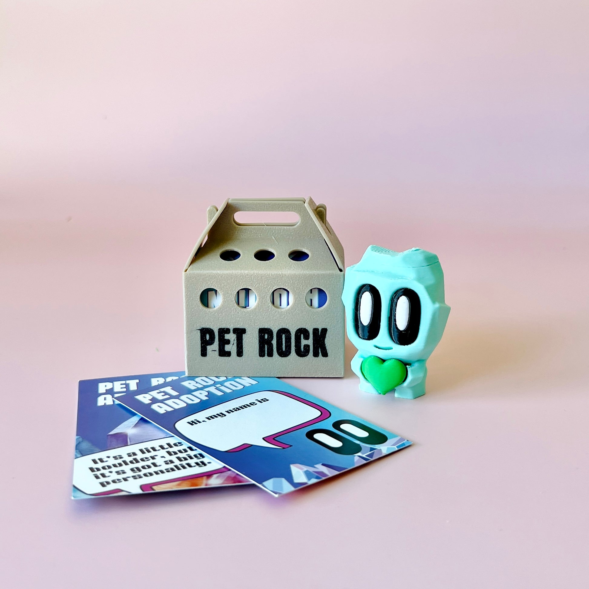 A quirky light blue figure with large eyes and a heart, alongside a gray box labeled "PET ROCK" and colorful adoption cards.