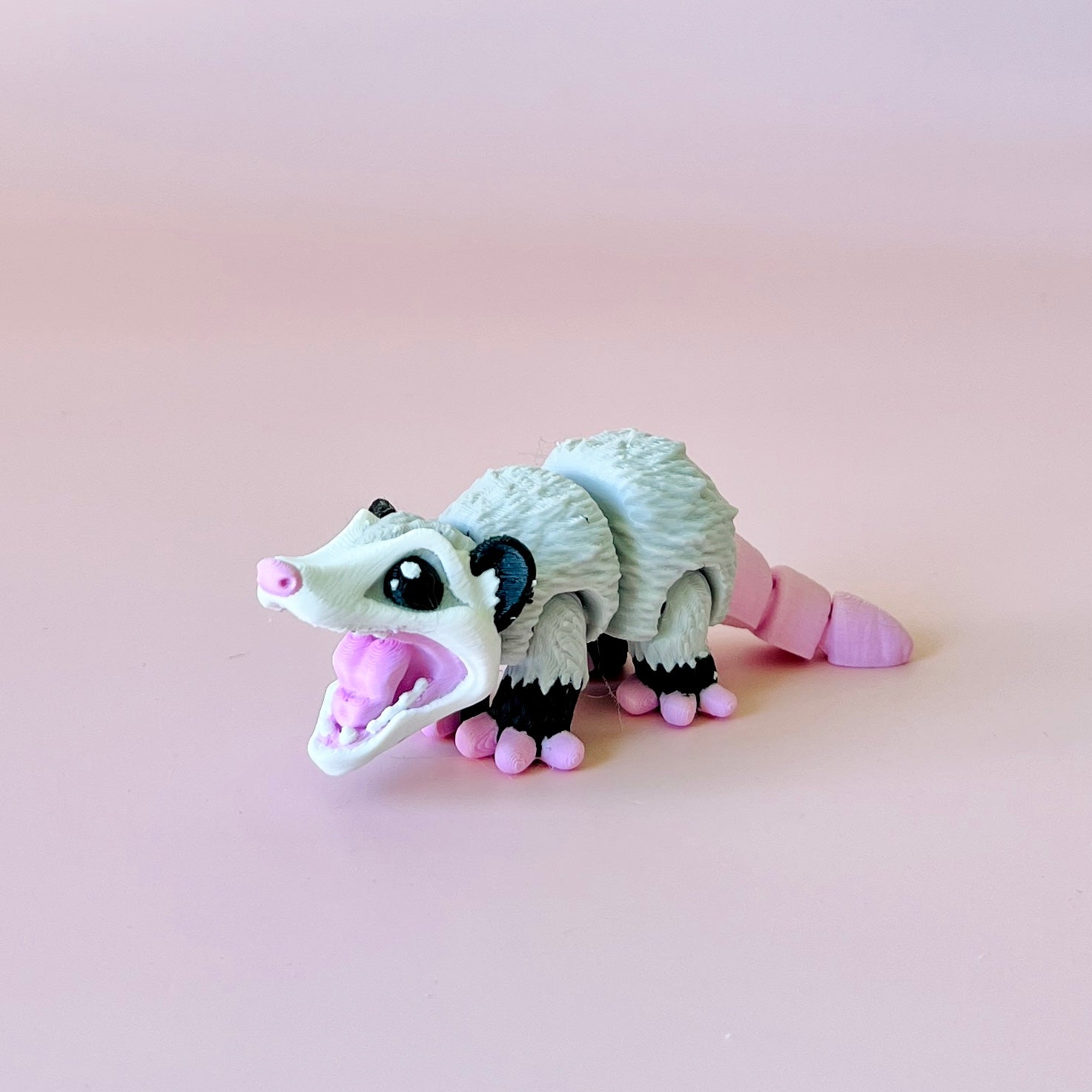A playful opossum figurine with a wide-open mouth, set against a soft pink background.