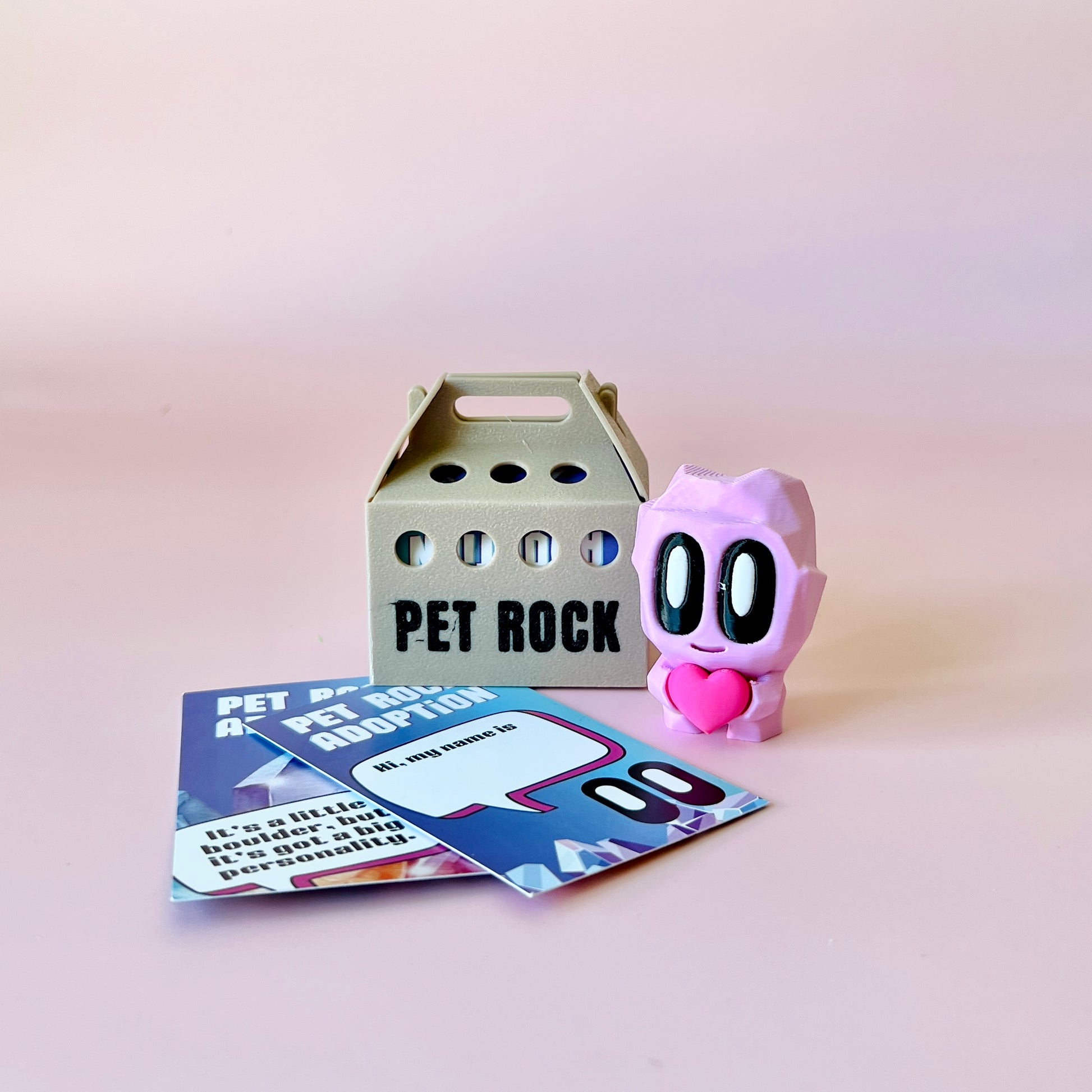 A cute pink character with big eyes and a heart stands beside a small carrier labeled "PET ROCK" and colorful cards.