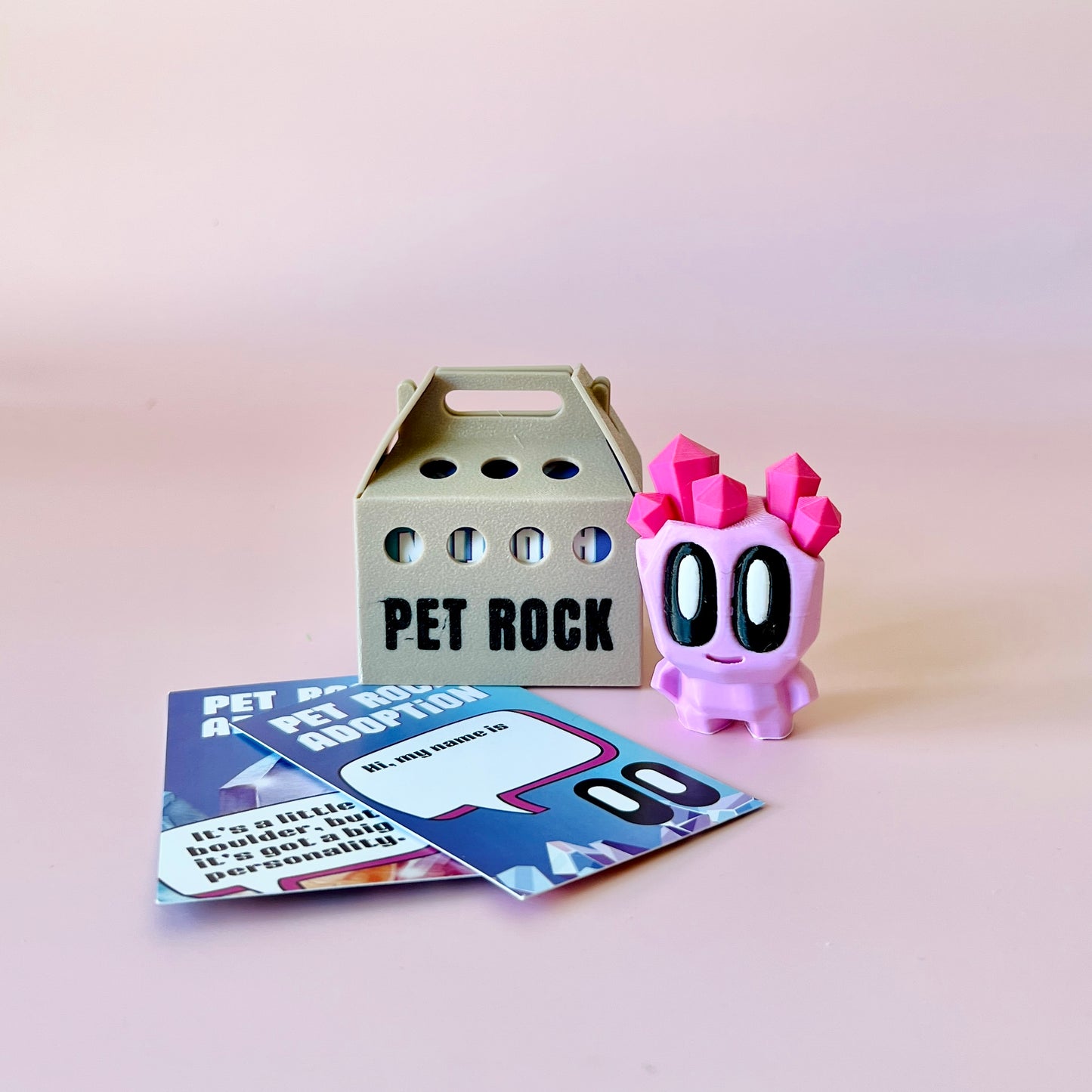 A playful pink character with crystal-like features beside a beige box labeled "PET ROCK" and colorful adoption cards.
