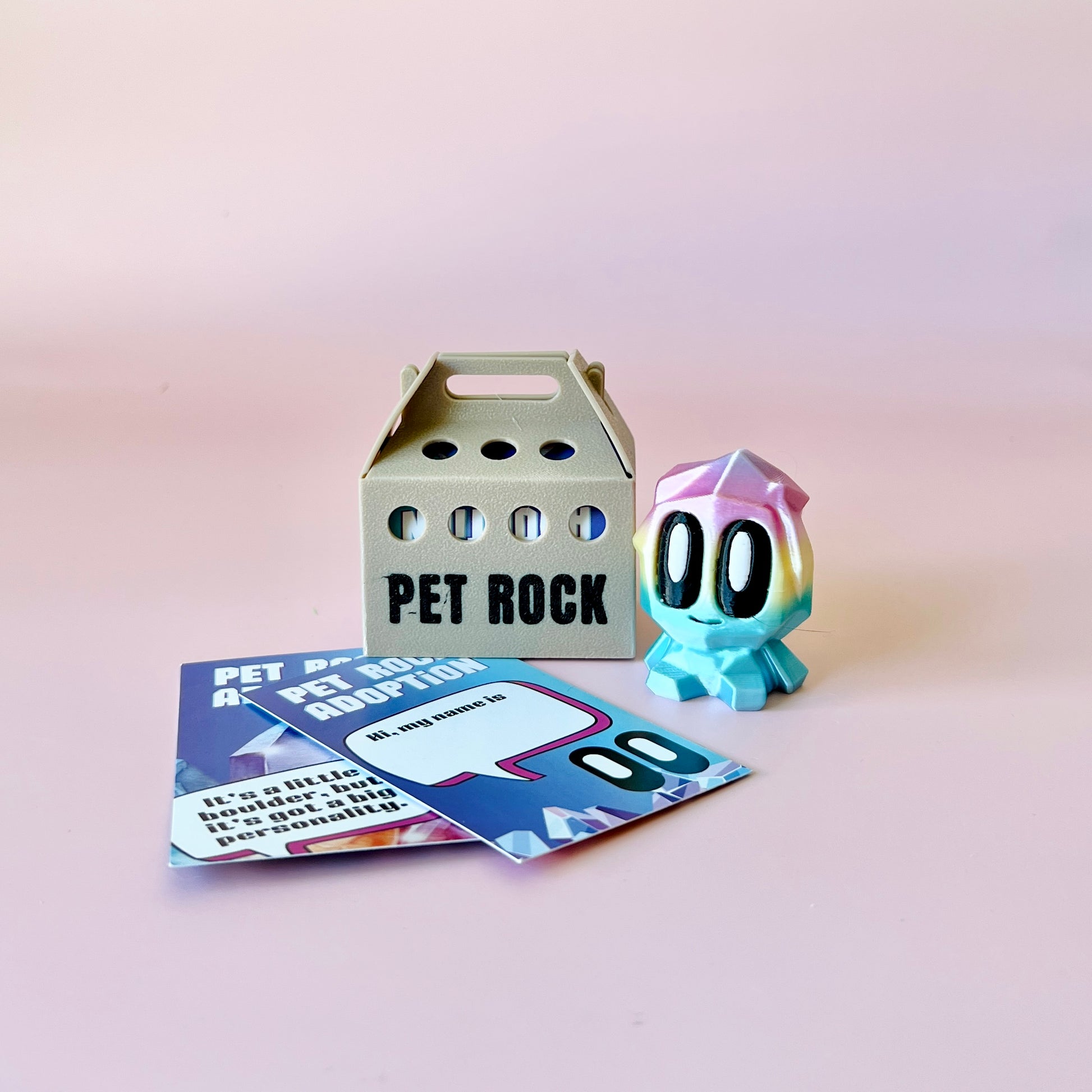A colorful, whimsical figure with large eyes beside a small carrier and adoption cards on a pink background.