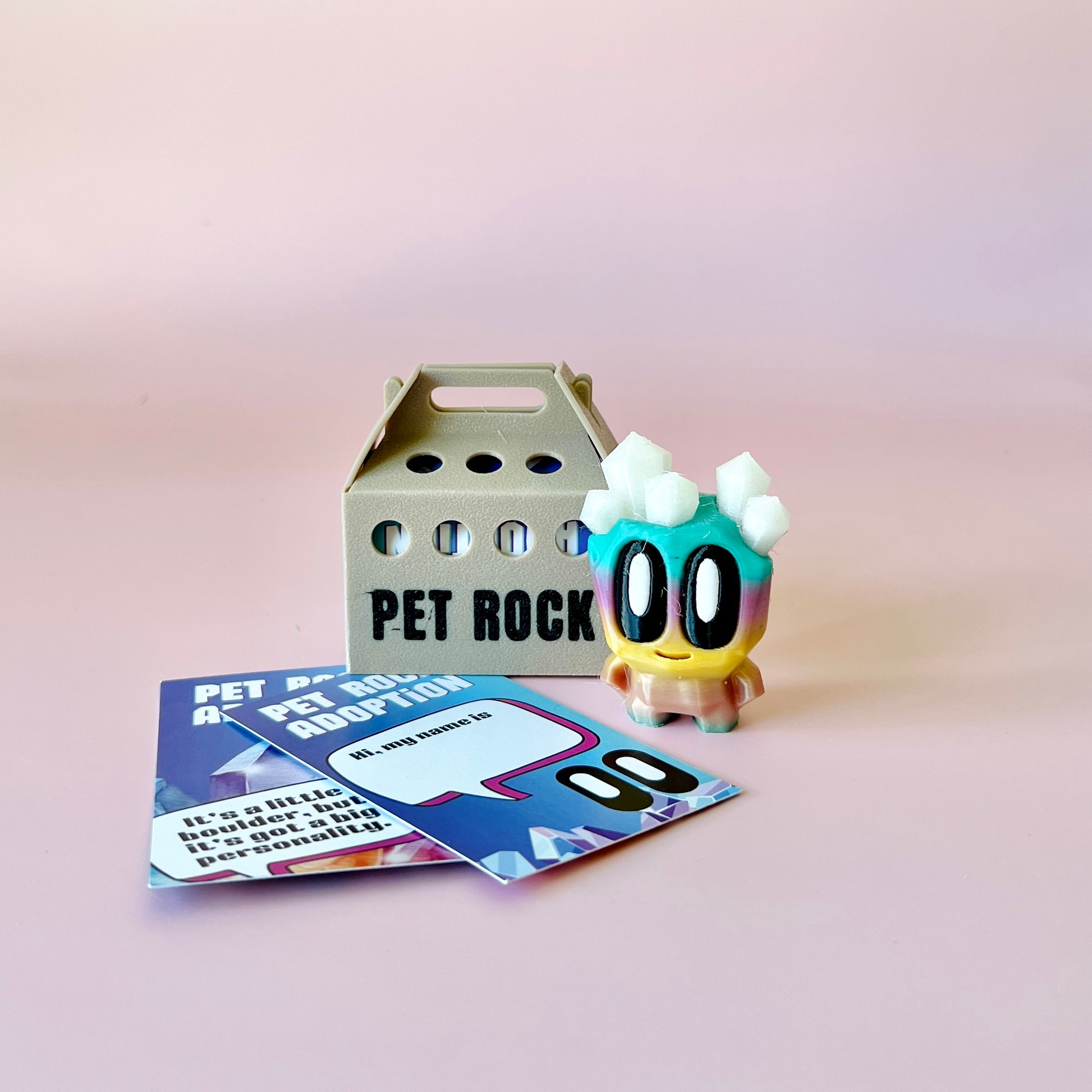 A colorful, cartoonish rock character with crystals on its head beside a gray pet rock carrier and adoption cards.