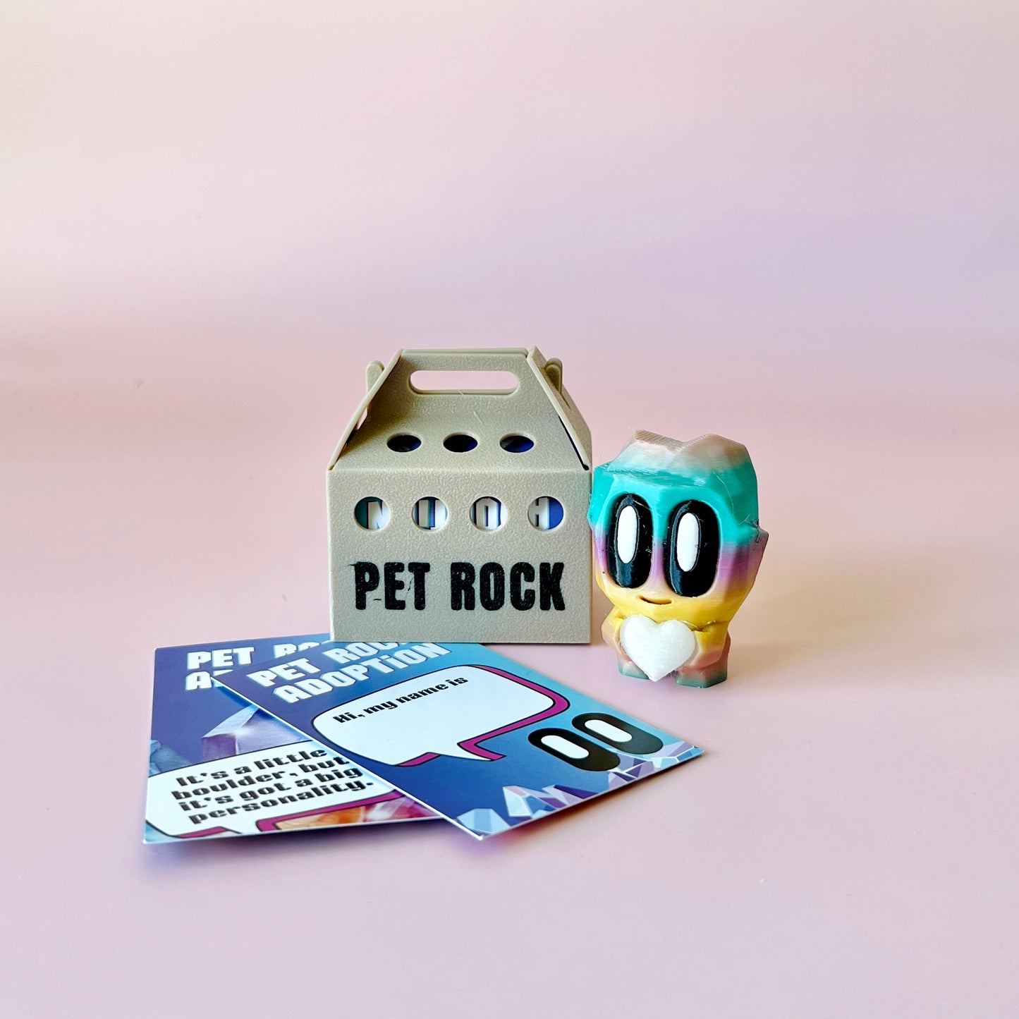 A colorful figure with big eyes and a heart stands beside a gray carrier labeled "PET ROCK" and adoption cards.