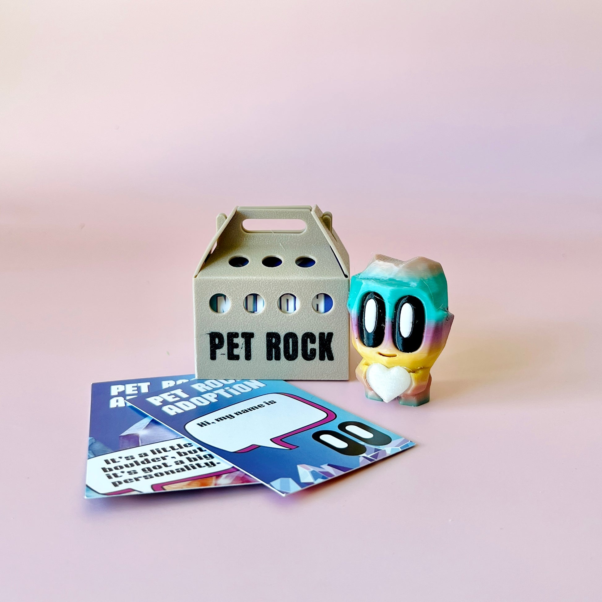 A colorful figure with big eyes and a heart stands beside a gray carrier labeled "PET ROCK" and adoption cards.